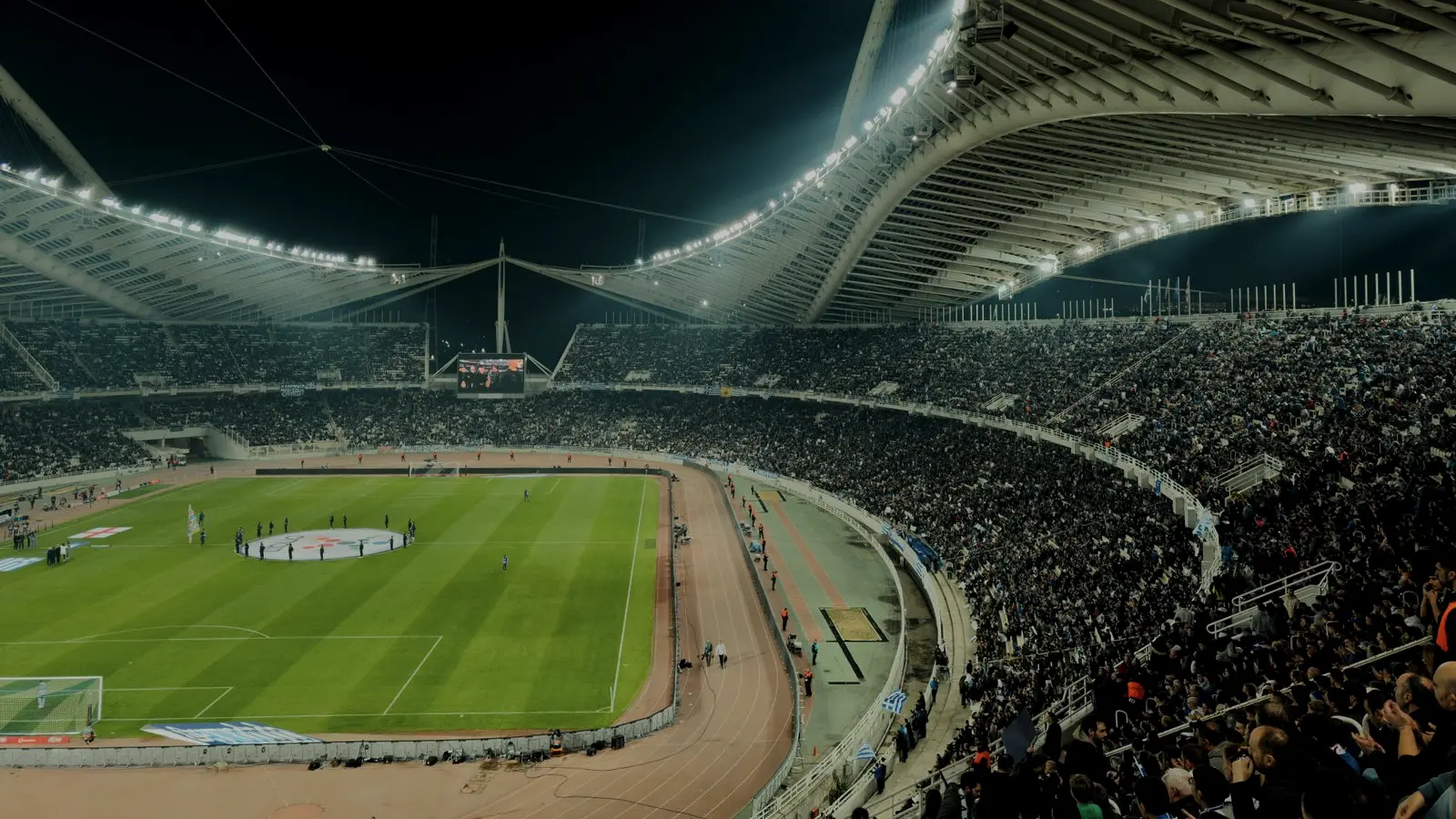 Estadio de fútbol iluminado por la noche durante un partido, con el público en las gradas.